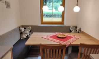 A cozy dining area with a wood-colored table and chairs. The room has a window with a view of greenery and is illuminated with warm light.