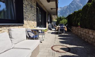 A cozy terrace with modern furniture and a view of the mountains. The surroundings are green and inviting.
