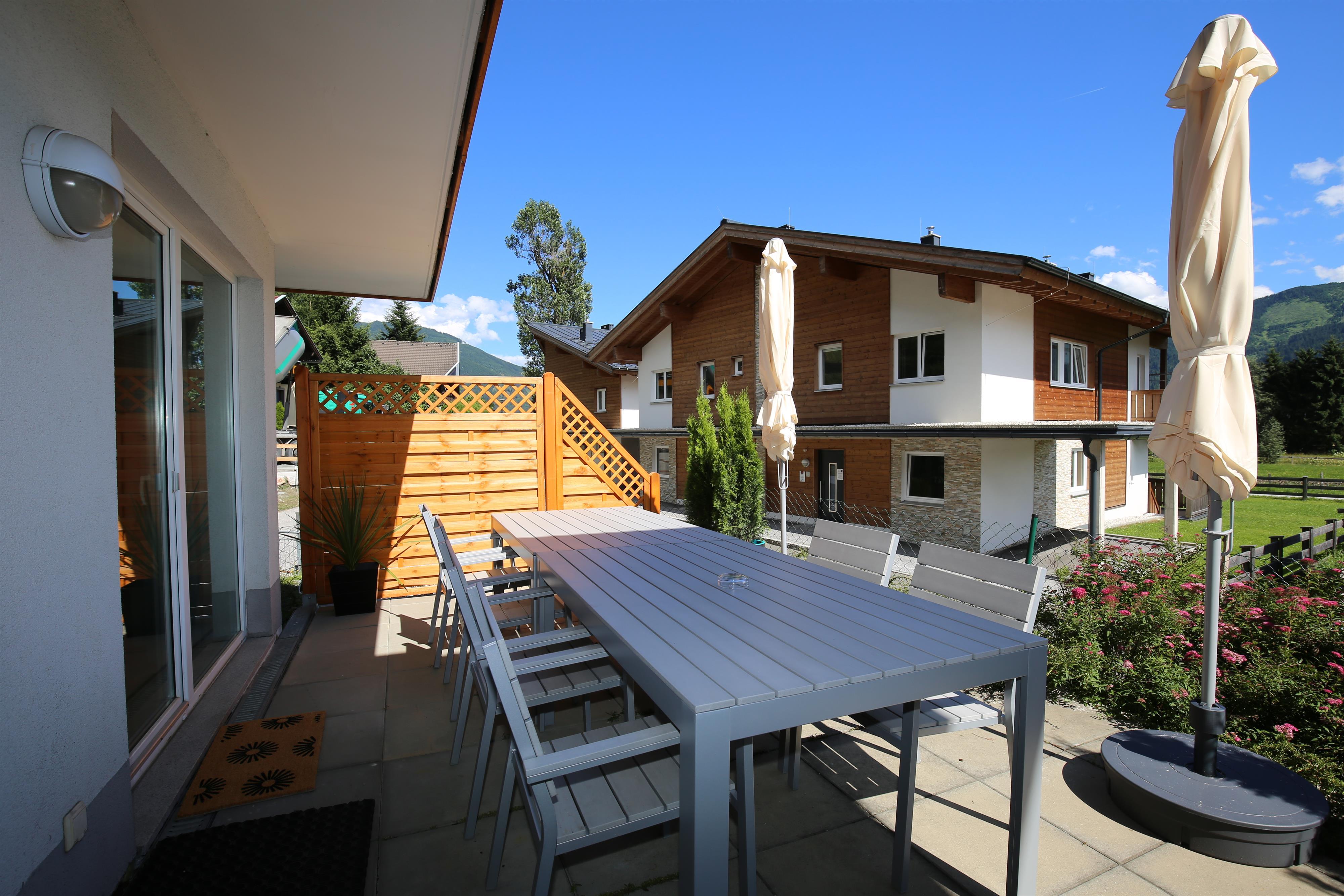 A beautiful outdoor area with a large table and chairs. In the background, typical alpine houses and a green landscape can be seen.