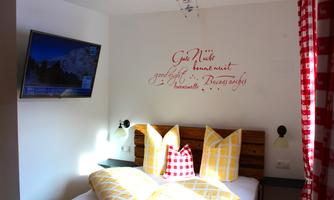 A cozy bedroom with a large bed and colorful cushions. There is a decorative writing on the wall, and a TV is mounted on the wall.