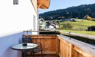 A cozy balcony with a table and a chair. In the background, green hills and typical houses are visible.