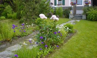 A well-kept garden with colorful flowers and a narrow stone path. In the background, a pretty house with shutters can be seen.