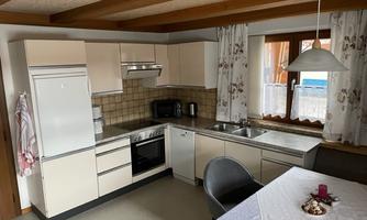 A modern kitchen with light cabinets and a dining area. Windows provide plenty of natural light and a view outside.