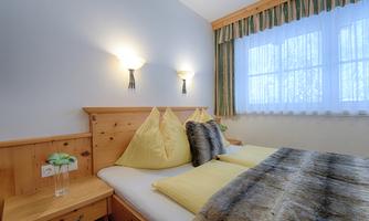 A cozy bedroom with a double bed and decorative pillows. The walls are light and the windows provide plenty of natural light.