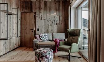 A cozy living space with wooden walls and a large window. The decor includes a gray sofa with colorful pillows, a green armchair, and a padded stool.