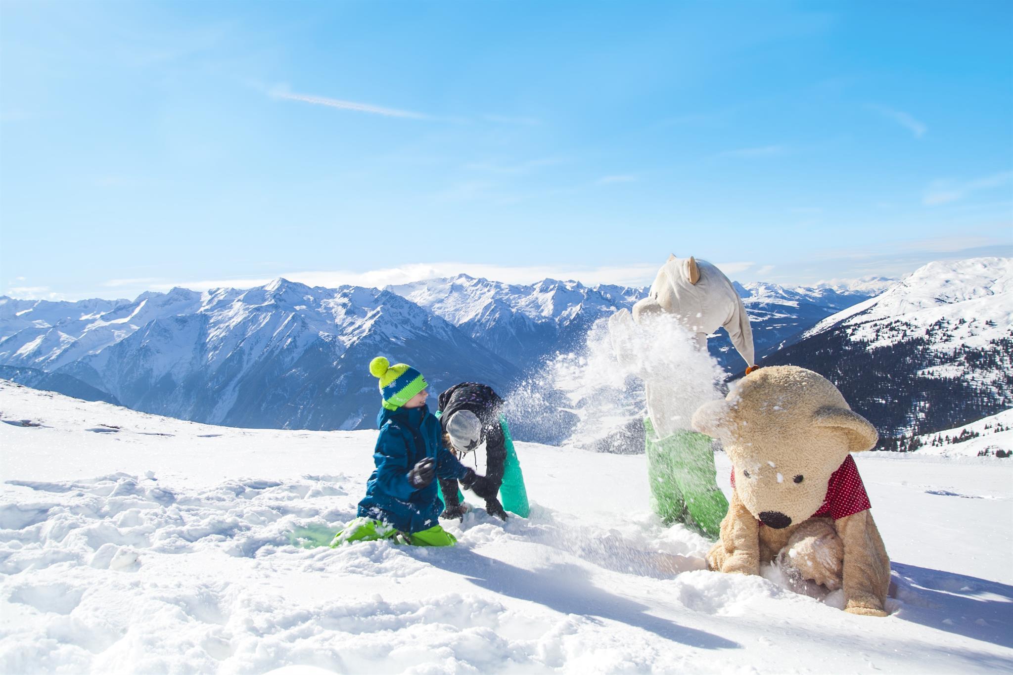 Children are playing in the snow with a large teddy bear. In the background, there are snow-covered mountains and a blue sky.