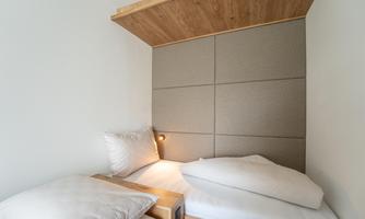 A simple room with two beds and bright, modern furnishings. The walls are light and there is cozy lighting.