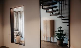 A modern interior with a wooden staircase and an elegant cabinet. On the left side, a large jug and curtains in the window are reflected.