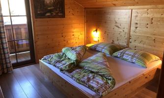 A cozy wooden bedroom with a large bed and colorful pillows. There is a window overlooking a balcony and warm light creates an inviting atmosphere.