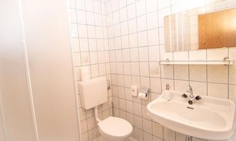 A simple bathroom with white tiles and a sink. The toilet is also present in the small, functional furnishings.