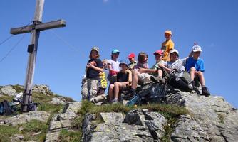 A group of children stands on a rock next to a wooden cross. The sky is clear and blue.