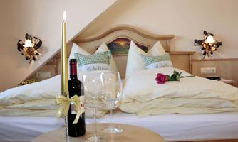 A cozy bedroom with a double bed and attractively decorated cushions. On the table are a bottle of wine, glasses, and a single rose.