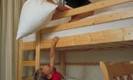 Two children are playing in a bedroom with bunk beds. The child on top is holding a pillow, while the child below is reaching for it.