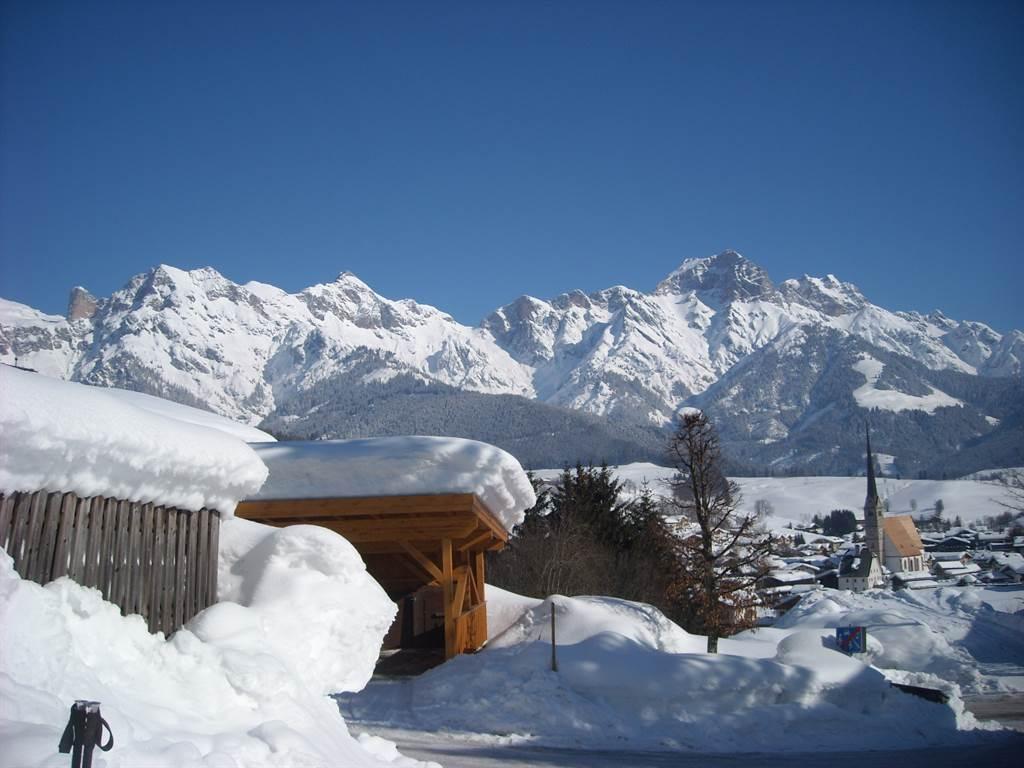 A snowy mountain landscape with majestic peaks in the background. In the foreground, a traditional building and snow-covered trees can be seen.