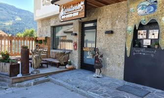 An inviting entrance to an accommodation with a wooden terrace and outdoor seating. Next to the door stands a decorative figure, and the surroundings are surrounded by mountains.