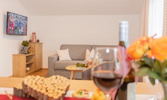 A cozy living room with a gray sofa, a wooden table, and a TV on the wall. On the table, there are wine and a vase of flowers that create an inviting atmosphere.