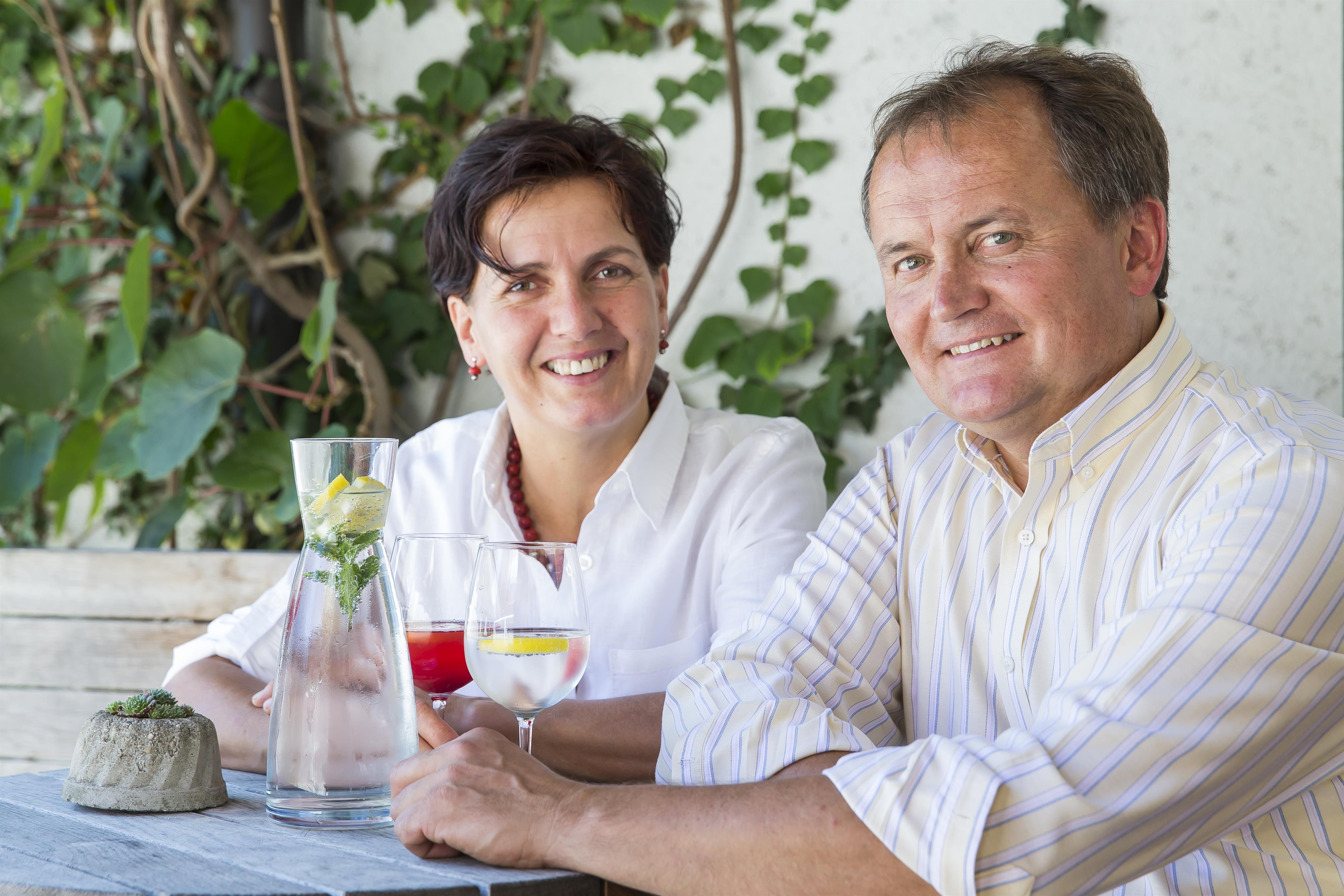 A friendly couple sits smiling at a table. They are enjoying drinks in a green, sunny environment.