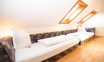 A simple attic room with two single beds and colorful sofas. The room has bright walls and windows that let in natural light.