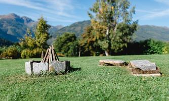 A campfire site with stones and wooden poles in a green meadow. In the background, trees and mountains are visible.