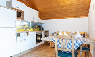 A modern kitchen with a wooden ceiling, white cabinets, and a dining table. On the table lies a striped tablecloth and glasses are prepared.