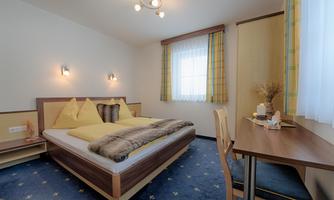 A cozy bedroom with a double bed and yellow pillows. A desk and bright windows create an inviting atmosphere.