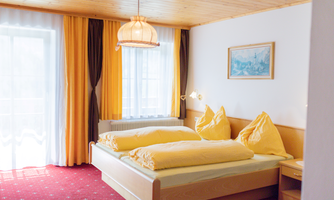 A bright bedroom with two beds and yellow bedding. Large windows with curtains let in plenty of light.