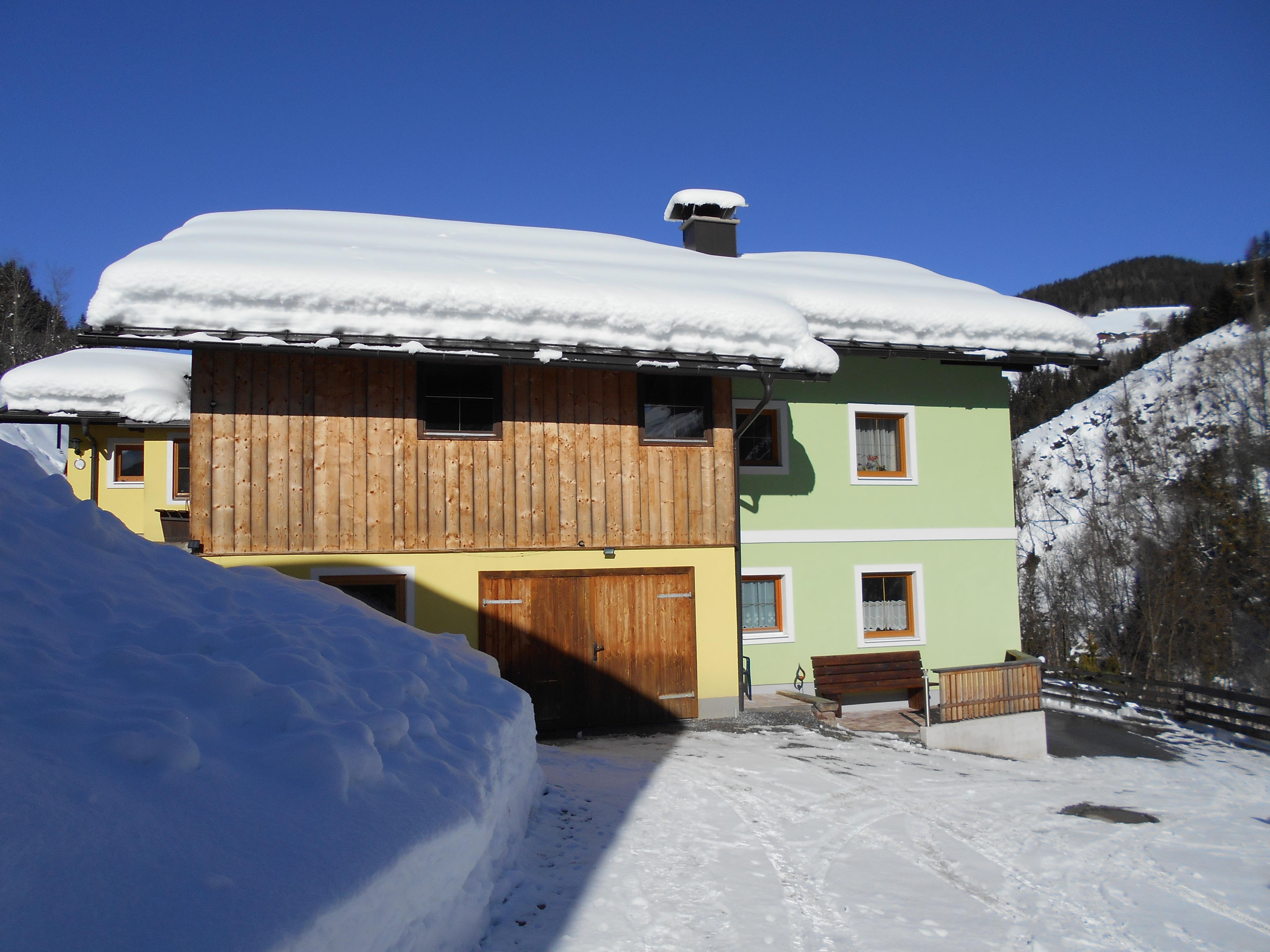 A charming, snow-covered house with a wooden and green facade. The surroundings are quiet and wintry with plenty of snow.