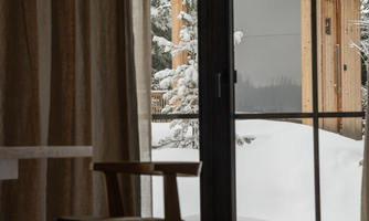 A cozy room with a wooden chair and a window overlooking a snowy landscape. Outside, a small, modern wooden cottage can be seen among the snow-covered trees.