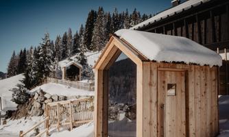 A wooden-clad cabin covered in snow stands in a winter landscape. In the background, snow-covered trees and another cabin can be seen.