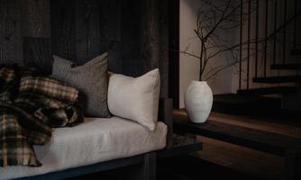 A cozy living room with a modern sofa, on which cushions and a blanket lie. Next to the sofa is a vase and in the background are stairs visible.