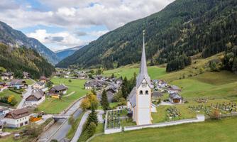 A picturesque village in the mountains with a striking church and surrounding green meadows. The landscape is characterized by gentle hills and a clear sky.