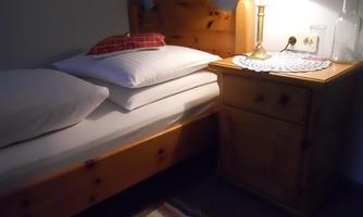 A simple bedroom with a wooden bed and two large pillows. Next to the bed stands a nightstand with a lamp and a glass.