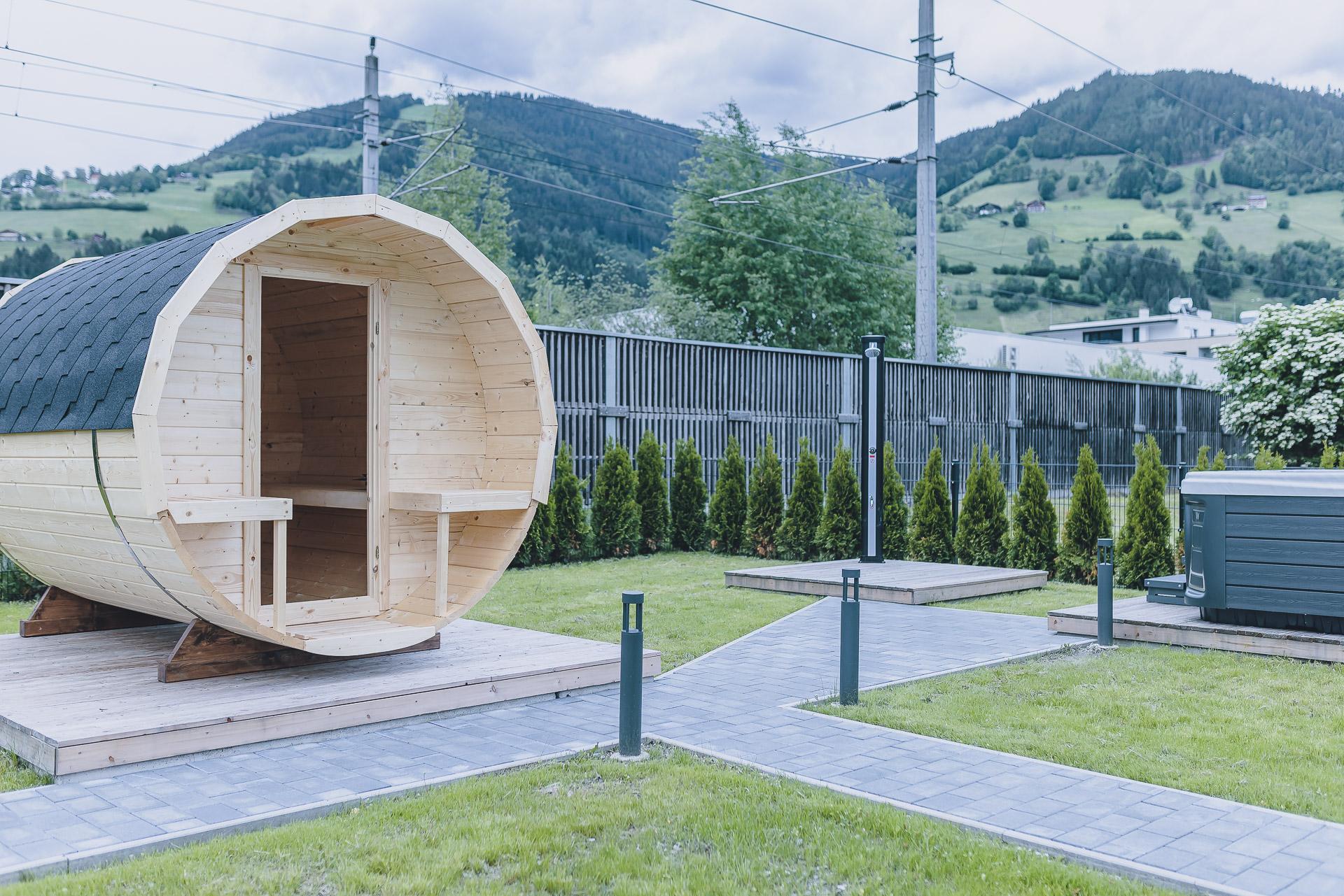 A cozy wooden cabin shaped like a barrel stands in a green garden. In the background, there are gentle hills and a clear sky.