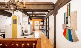 A bright hallway with wooden beams and modern artworks on the walls. On the left side, stylized walls and an open kitchen are visible.