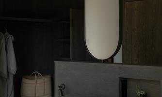 A modern bedroom with a large mirror and white bedding. The walls are in dark tones and the light is soft and inviting.