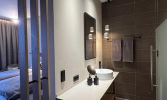 A modern bathroom with an elegant sink setup and large mirrors. In the background, a bedroom with a cozy bed is visible.