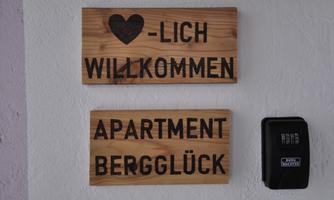 A beautiful wooden welcome sign with the inscription "Warm Welcome" and "Apartment Bergglück." Next to it is a black key box.