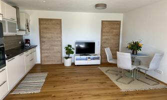 A modern apartment with an open kitchen and a living room. There is a dining table with chairs and a television on a dresser.