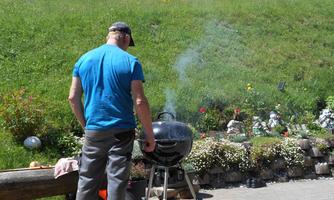 A man is grilling outdoors in a green, blooming landscape. In the background, decorative figures and flowers can be seen.
