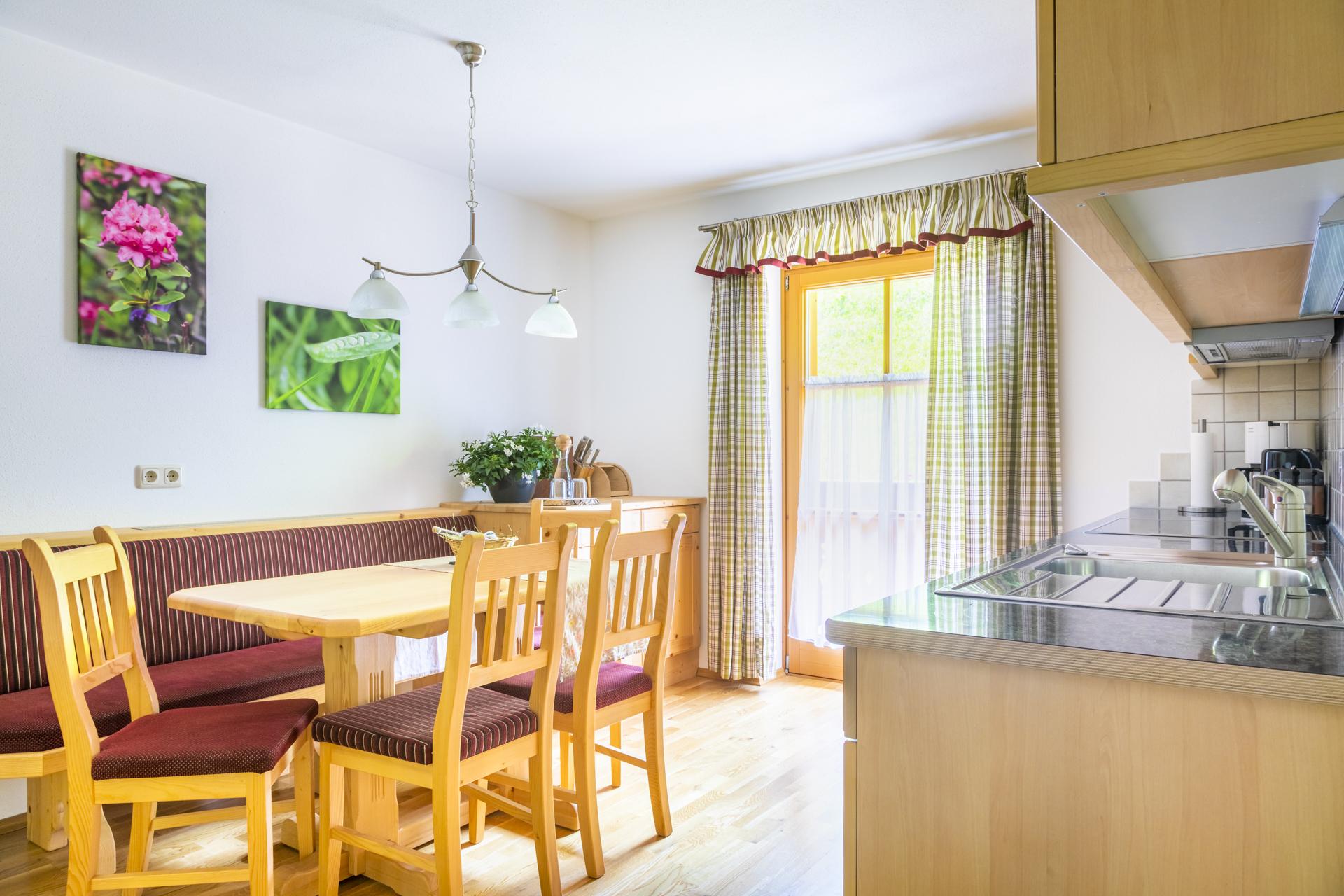 A bright kitchen with a wooden dining table and chairs. Pictures of plants hang on the walls, and the windows are decorated with a curtain.