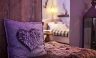 A cozy room with a purple pillow featuring a heart motif. Soft textiles and warm light can be seen in the background.