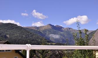 A panoramic view of the mountains under a clear blue sky. In the foreground, there is a balcony with a railing and some green foliage.