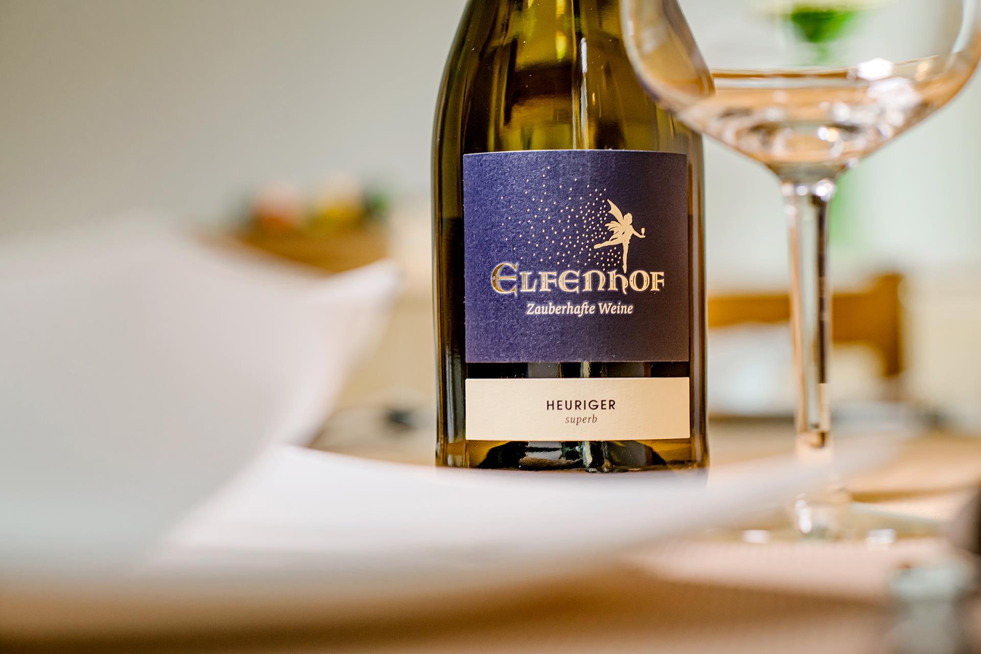 A bottle of Elfenhof wine stands on a table, surrounded by glassware and cutlery. The style is simple and elegant.