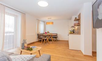 A modern living room with wooden flooring and cozy furnishings. There is a dining area and plenty of natural light.