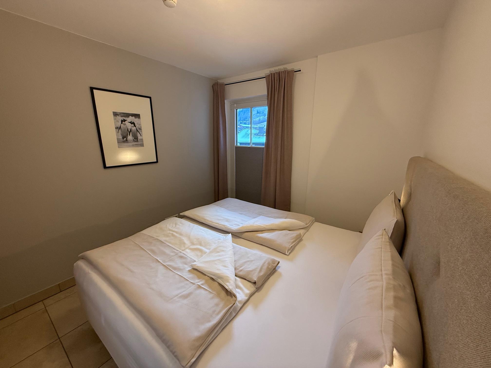 A cozy bedroom with a double bed and two single blankets. The walls are painted in neutral colors and a window provides daylight.