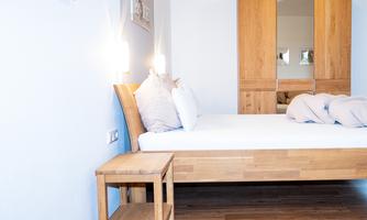 A cozy bedroom with a wooden bed and a nightstand. The walls are bright and there is a large wardrobe.