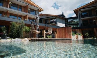 An elegant hotel with a clear pool in the foreground. Hanging chairs and green plants create a relaxed atmosphere.