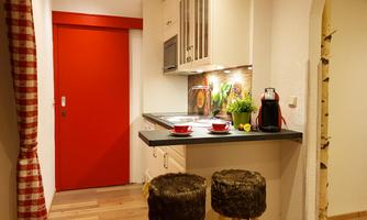 A modern kitchen with a red door and wooden floor. Two stools and a coffee machine complete the space.