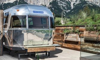 A modern caravan sits on a terrace overlooking impressive mountains. The surroundings are green and inviting, perfect for relaxing.