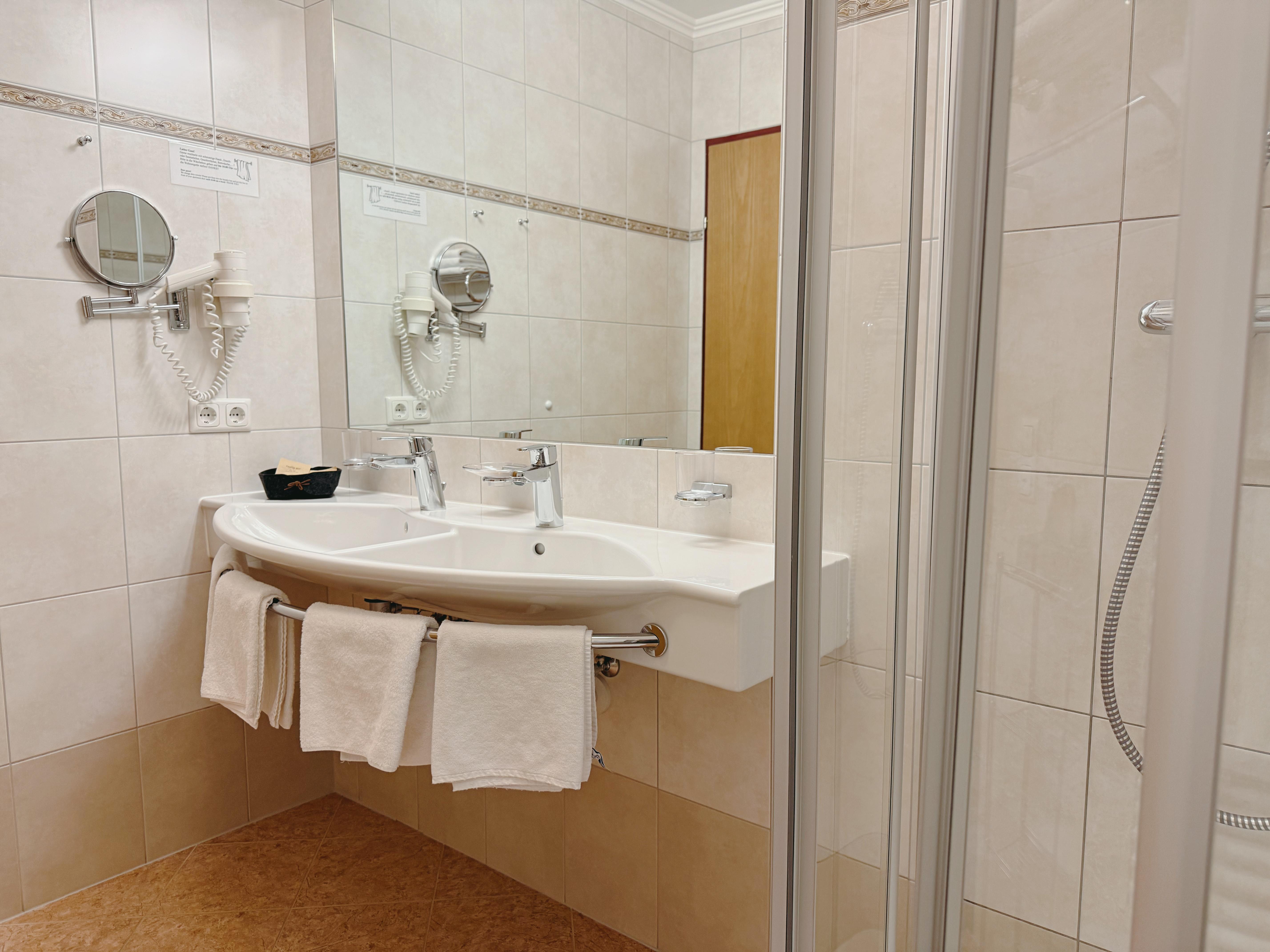 A modern bathroom with a double sink unit and towels. The walls are brightly tiled, and there is a shower in the background.